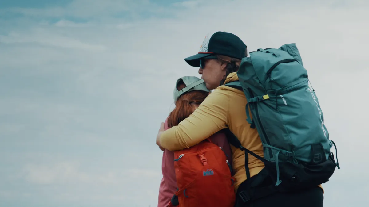 Two hikers with large packs hug on a ridge under a pale sky.
